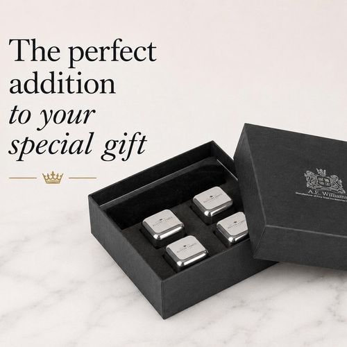 A set of four polished whiskey chilling cubes with rounded edges, neatly arranged inside a black presentation box. Each cube is engraved on top with the "A E WILLIAMS" crown logo. The open gift box rests on a light-coloured marble surface next to its matching black lid, which features a silver crest emblem and text reading A.E. Williams