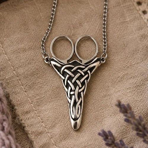 A pewter chatelaine scissor sheath featuring an intricate Celtic knotwork design, hanging from a metal chain with small scissors inserted, set against a rustic linen background.