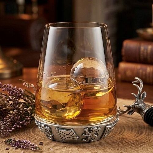 A curved glass whiskey tumbler filled with golden scotch and two rounded ice spheres, featuring a heavy pewter base intricately detailed with an embossed pattern of Scottish thistles, a rampant lion, and the Saltire flag