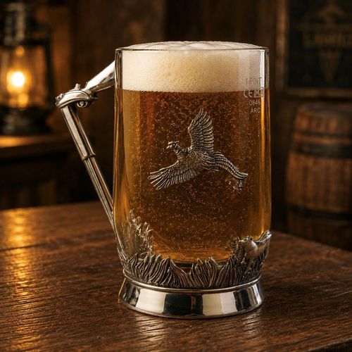 A clear glass beer tankard filled with golden, bubbling lager and a thick white foam head, resting on a rustic wooden table. The glass features an angular pewter handle shaped like a detailed shotgun, with the barrel extending downwards. The front of the glass is decorated with a detailed pewter relief of a pheasant in mid-flight, while the base wrap depicts a countryside hunting scene with thick reeds, marsh grass, and a hunting dog giving chase on the right