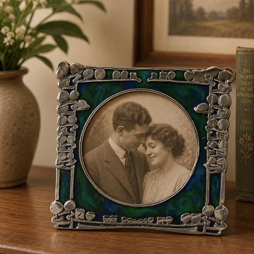 An Art Nouveau square pewter picture frame featuring an intricate honest plant design, a vibrant blue-green enamel backdrop, and a circular opening displaying a vintage Edwardian couple's portrait