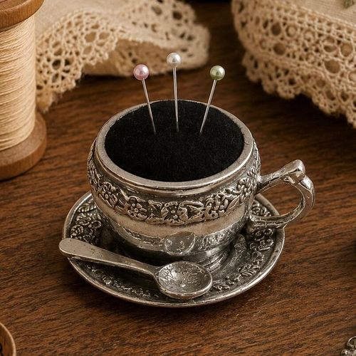 Vintage-style ornate pewter teacup pincushion on a matching saucer with three pearl-head pins stuck in the black cushion