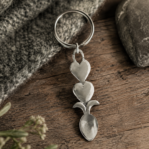 Handcrafted pewter Welsh lovespoon keyring featuring two stacked smooth hearts above a spoon bowl, set on a rustic wooden surface next to grey knit fabric and a smooth stone.