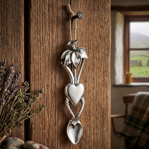 Handcrafted pewter Welsh lovespoon hanging from a twine loop on a rustic wooden beam, featuring a top daffodil flower motif, a smooth solid heart center, a twisted rope pattern, and a textured heart-shaped spoon bowl.