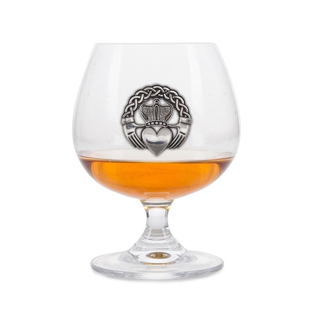 Crystal brandy glass with pewter Claddagh emblem, partially filled with amber liquid
