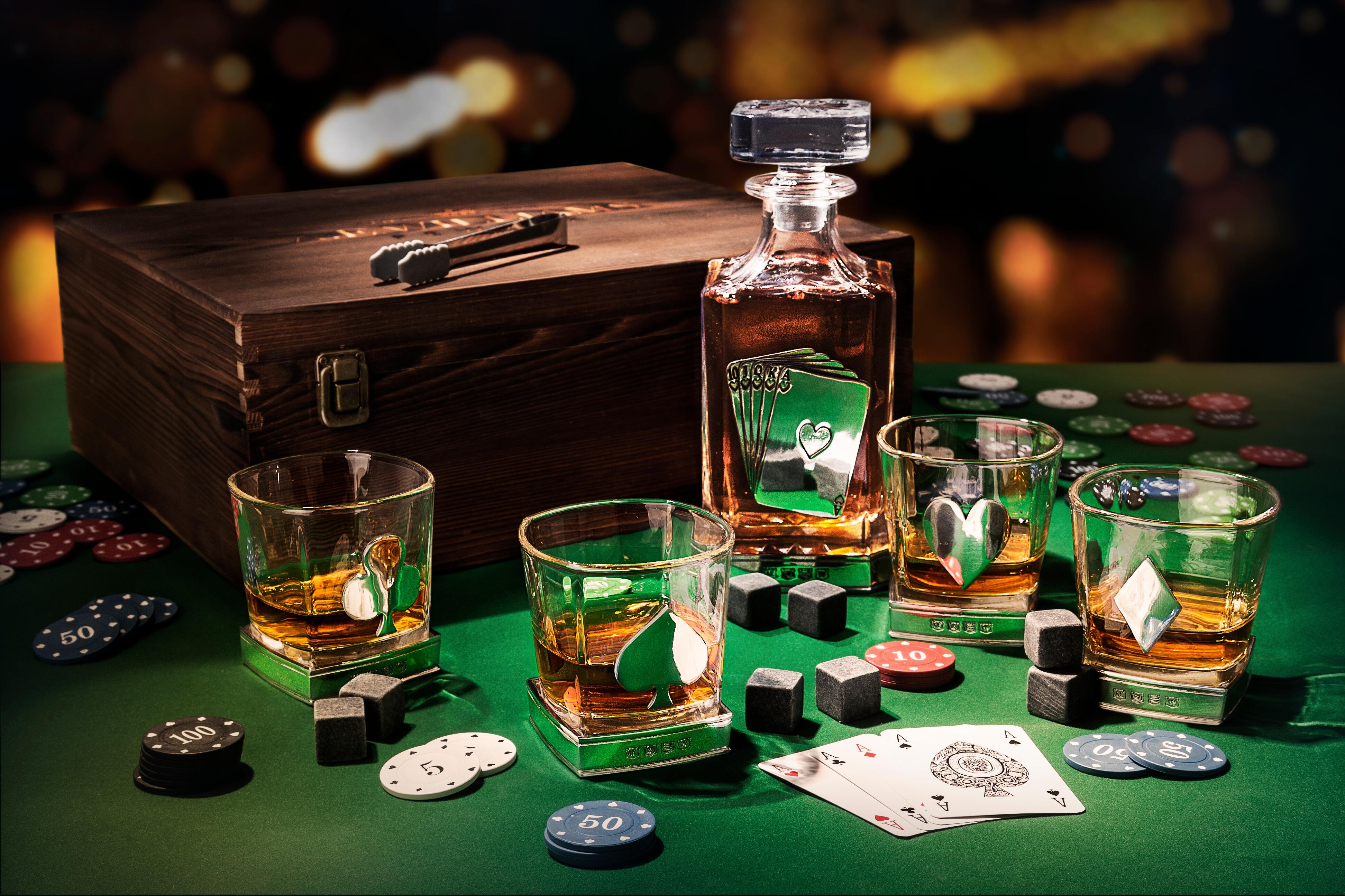 Whiskey decanter set with playing card theme arranged on a green poker table with playing cards, poker chips, and whiskey stones scattered around. The wooden box and glasses feature card suit designs, filled with amber whiskey