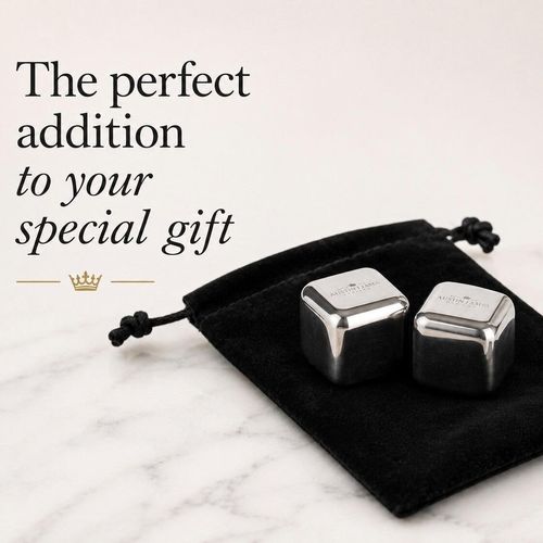 Two polished whiskey chilling cubes with rounded edges, resting on a small black velvet drawstring pouch. The top surface of each silver-coloured metal cube is subtly engraved with a brand logo reading "A E WILLIAMS" under a small crown