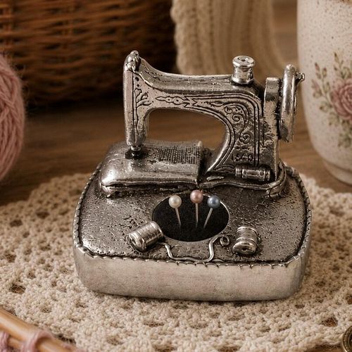 A miniature vintage sewing machine pin cushion made of detailed pewter, featuring a small black velvet cushion insert, molded thread spools, and pearl-headed pins, resting on a textured knit background.