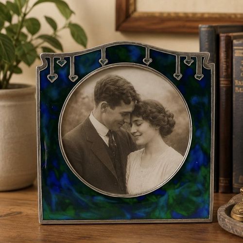 An Art Nouveau square pewter picture frame featuring an intricate honest plant design, a vibrant blue-green enamel backdrop, and a circular opening displaying a vintage Edwardian couple's portrait