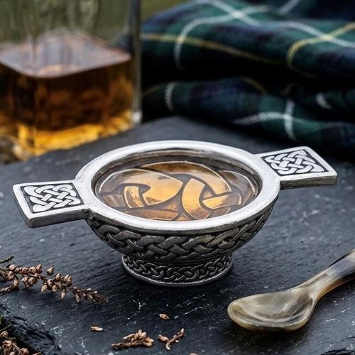 Pewter quaich bowl filled with whiskey, featuring rectangular handles and an exterior intricately embossed with Celtic interlace knotwork, displayed on slate beside a horn spoon and tartan cloth