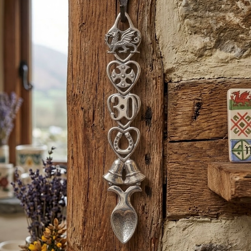 Handcrafted pewter 30th anniversary Welsh lovespoon hanging on a rustic wooden beam, featuring detailed symbols including a Welsh dragon, Celtic cross, the number 30, a horseshoe, wedding bells, and a heart-shaped spoon bowl.