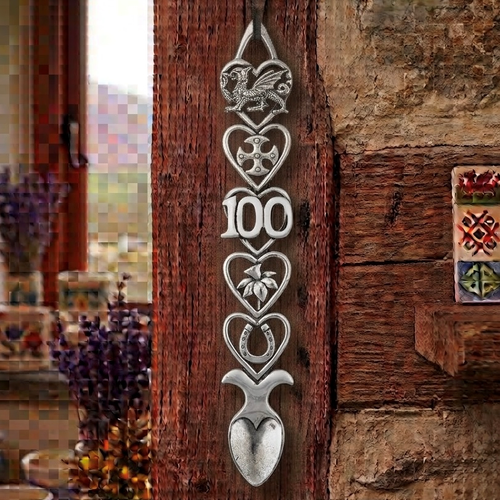 Handcrafted pewter 100th birthday Welsh lovespoon hanging on a rustic wooden beam, featuring detailed symbols including a Welsh dragon, Celtic cross, the number 100, a daffodil, a horseshoe, and a heart-shaped spoon bowl.