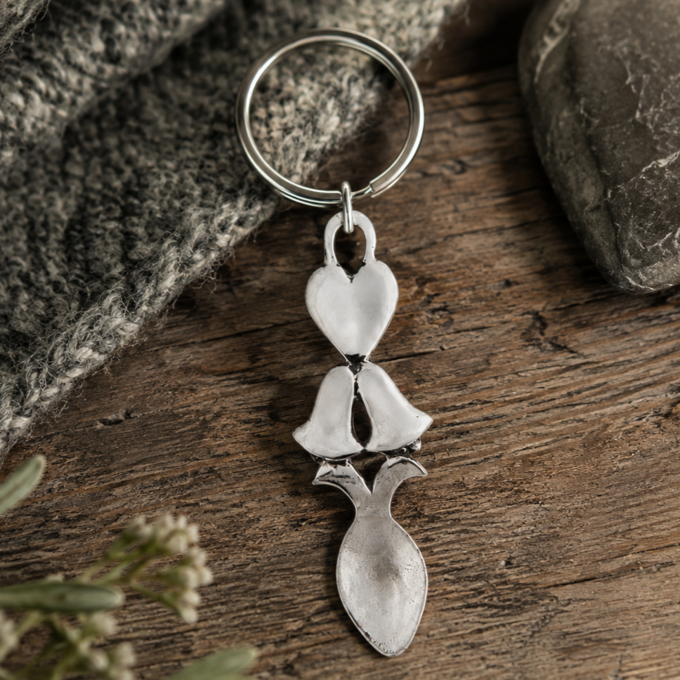 Handcrafted pewter Welsh lovespoon keyring featuring a smooth heart motif, wedding bells, and a spoon bowl, resting on a rustic wooden surface next to grey knit fabric and a smooth stone.