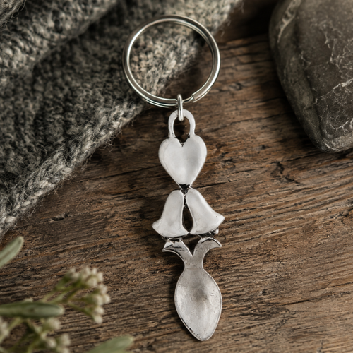 Handcrafted pewter Welsh lovespoon keyring featuring a smooth heart motif, wedding bells, and a spoon bowl, resting on a rustic wooden surface next to grey knit fabric and a smooth stone.