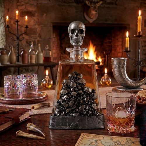 A clear glass spirits decanter filled with amber liquid, sitting in a dark, stone-textured pewter base. The front of the decanter is adorned with a large, highly detailed pewter relief depicting a piled pyramid of small human skulls. The bottle is sealed with a glass stopper topped by a prominent, sculpted pewter skull