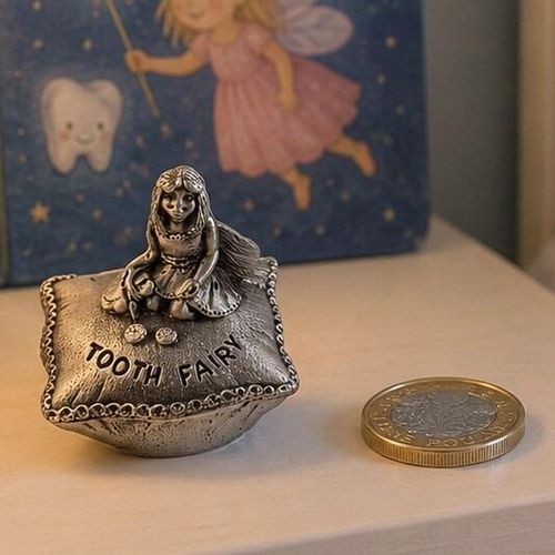 A small, detailed pewter keepsake box shaped like a tufted pillow or cushion, displayed next to a British one pound coin for scale on a light wooden surface. The top of the cushion is engraved with the words "TOOTH FAIRY" and features a sculpted miniature fairy figure kneeling with long hair and delicate wings. The box lid has an ornamental, patterned trim along the edge