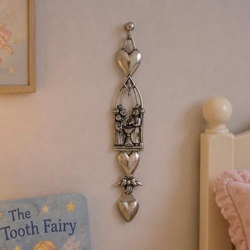 An elongated, decorative pewter Welsh lovespoon hanging vertically from a metal peg on a plain, light-colored wall. The intricate lovespoon features a polished heart at the top, followed by an openwork Gothic arch frame enclosing a sculpted relief scene of a christening or baptism over a font bowl. Below the scene is a second smooth heart, a small decorative ribbon bow, and a textured, heart-shaped spoon bowl at the bottom