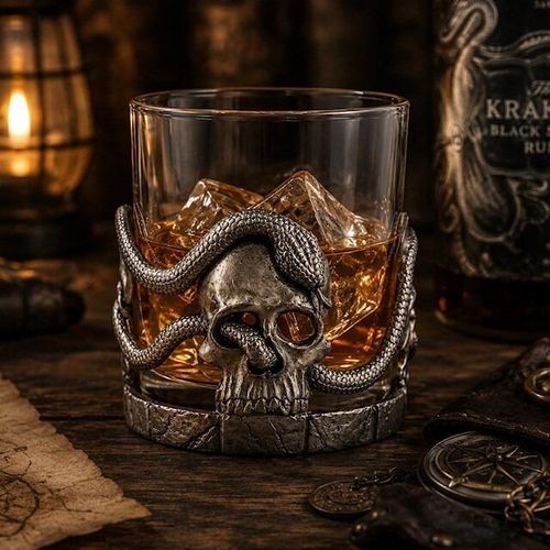 A lowball glass whisky tumbler filled with dark amber spirits and ice cubes, encased in a dramatic, gothic-style pewter holder. The front of the holder features a prominent, high-relief human skull with a detailed textured serpent or snake wrapping around it, slithering across the brow and emerging through one of the eye sockets