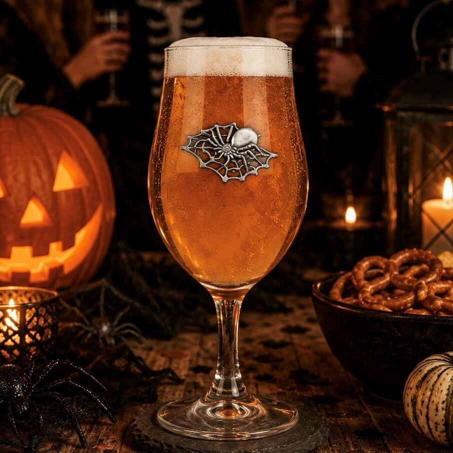 A stemmed beer glass filled with amber ale and a frothy head, featuring a detailed pewter spider on a web emblem on the front