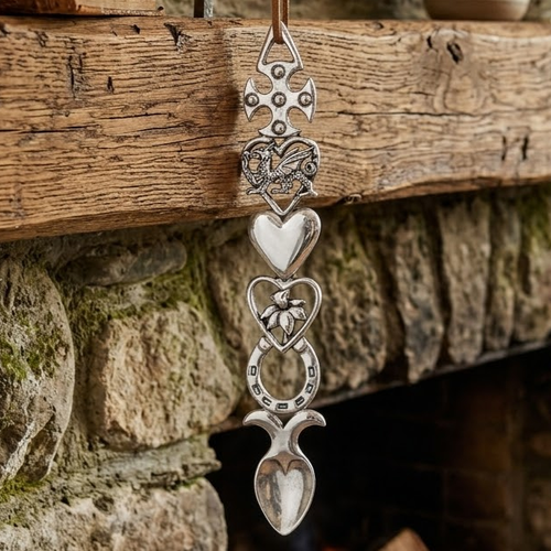 Handcrafted pewter Welsh lovespoon hanging against a wooden beam and stone fireplace, featuring a studded Celtic cross top, a dragon inside an open heart, a smooth solid heart, a daffodil inside an open heart, a lucky horseshoe, and a spoon bowl.