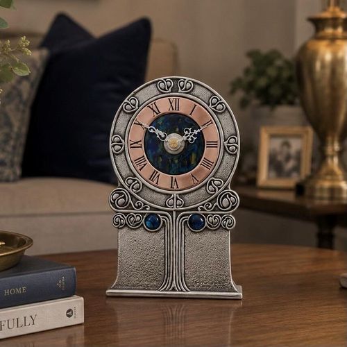 An Arts and Crafts style pewter mantel clock designed by Archibald Knox, featuring a circular upper frame and a textured rectangular base with an embossed stylized tree design. The clock face has a copper chapter ring with Roman numerals surrounding a deep blue and green enamelled centre, bordered by repeating Celtic knot motifs and accented with two dark blue cabochon gems below the dial.