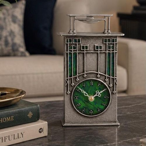 An Arts and Crafts style pewter carriage clock designed by Archibald Knox, featuring a tall, textured rectangular frame topped with a flat lid and a stylized overhead handle. A circular clock face with a swirling green enamelled background and decorative pewter lettering is positioned near the bottom, beneath geometric Art Nouveau details inlaid with green and blue enamel.