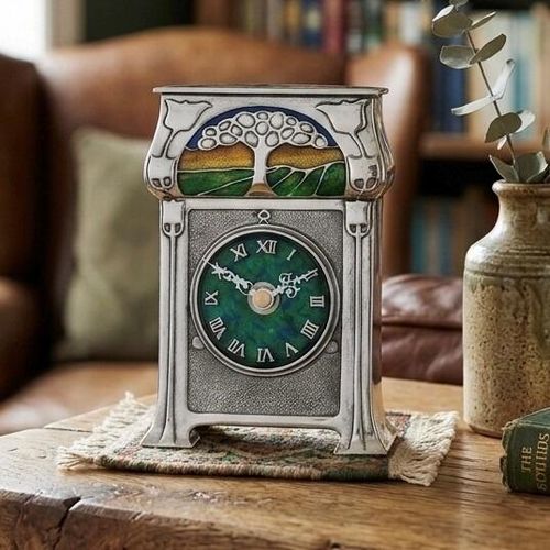 An Arts and Crafts style pewter mantel clock designed by Archibald Knox, featuring a vibrant green and blue enamelled clock face with Roman numerals. The top of the clock is adorned with an enamelled landscape design of a stylized tree under a blue sky