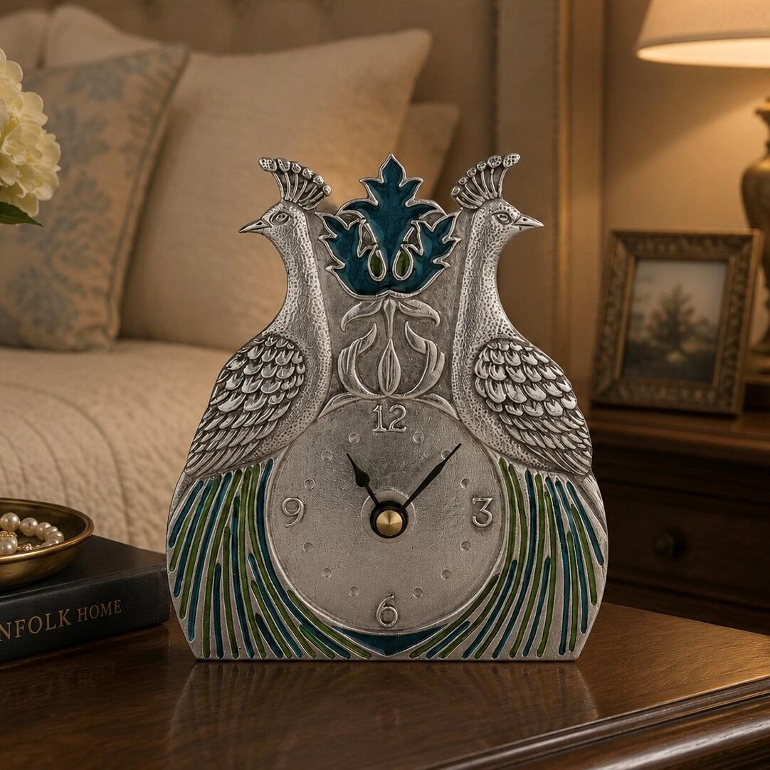 An Arts and Crafts style pewter desk clock inspired by William Morris, featuring a symmetrical design with two embossed peacocks facing outward on either side of the frame. The textured pewter-toned clock face displays large numbers for 12, 3, 6, and 9, and is framed at the bottom by flowing peacock feathers inlaid with green and blue enamel, with an ornate blue floral motif at the top
