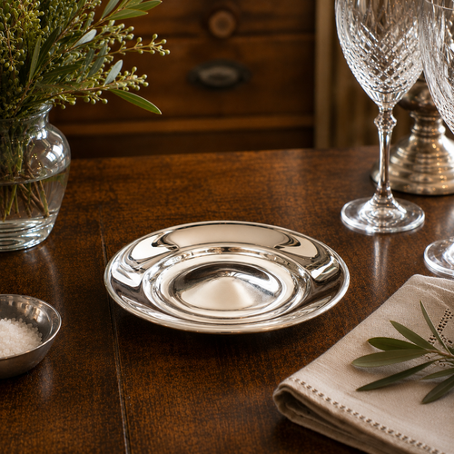 Handcrafted pewter 9 inch plate displayed in an elegant dining setting with crystal glassware and greenery, featuring a bright mirror-polished finish and raised centre detail.