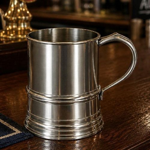 A classic one-pint pewter tankard with a brushed metallic finish and traditional beaded raised bands around its lower half. The tankard features an elegant curved handle and sits prominently on a dark wooden bar counter in a traditional British pub