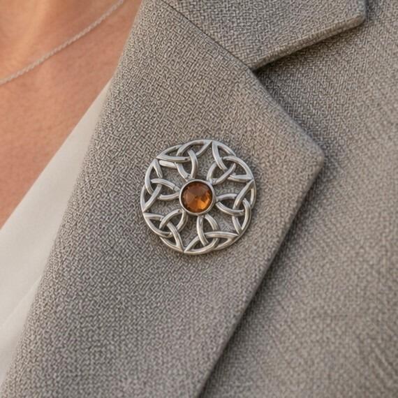 A circular pewter brooch featuring a symmetrical openwork Celtic knot design and a central faceted amber-coloured stone