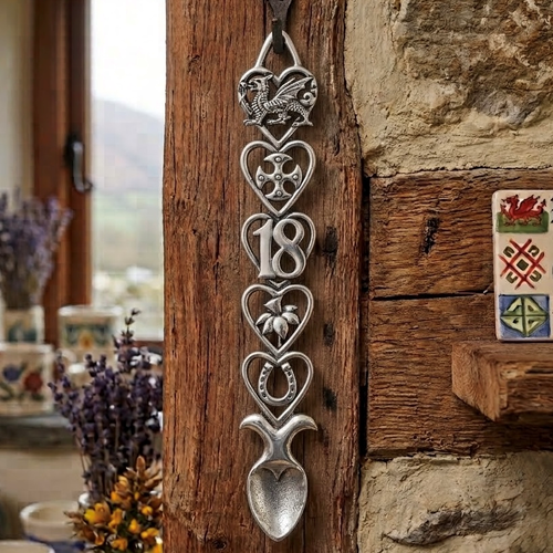 Handcrafted pewter 18th birthday Welsh lovespoon hanging on a rustic wooden beam, featuring detailed symbols including a Welsh dragon, Celtic cross, the number 18, a daffodil, a horseshoe, and a heart-shaped spoon bowl.