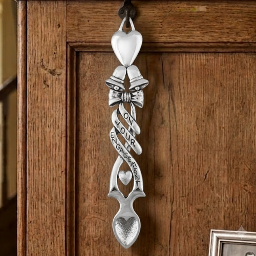 Handcrafted pewter Welsh lovespoon hanging on a wrought iron hook against a wooden panel, featuring a smooth solid top heart, twin wedding bells with a bow, a ribbon engraved with "ON YOUR ENGAGEMENT", a small suspended heart, and a textured spoon bowl.