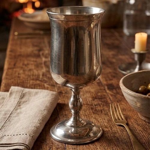 A traditional tulip-style pewter goblet with a smooth, brushed metallic finish. The goblet features an elegant bell or tulip-shaped bowl with a softly flared rim, supported by a classic baluster stem with a central spherical detail and a wide, stepped circular base