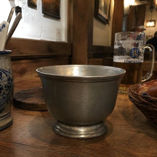 A traditional Bavarian-style pewter bowl or chalice with a brushed, matte metallic finish and an antique patina. The vessel features a deep, rounded bowl with a slightly flared rim, supported by a short, sturdy pedestal base with stepped circular detailing. It sits on a rustic wooden table inside a German tavern, with a Löwenbräu beer stein