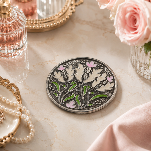 William Morris-inspired pewter purse mirror featuring cream floral motifs with green leaves and pink accents, displayed on an elegant vanity beside roses, pearls, and perfume.