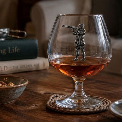 A short-stemmed clear glass brandy snifter filled with amber brandy, resting on a woven rope coaster on a rustic wooden table. The front of the glass bowl is adorned with a detailed pewter relief emblem of a shooter or hunter in traditional country attire, standing and aiming a shotgun upward
