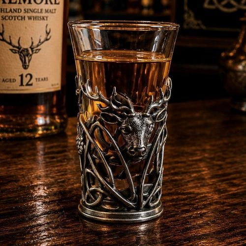 A tall glass shot glass filled with amber-colored liquor, sitting on a dark wooden bar counter. The lower half of the glass is encased in an intricate pewter metal holder featuring an openwork Celtic knot and foliage design. A highly detailed, relief-sculpted head of a Highland stag with large, prominent antlers is centred on the front of the metal holder