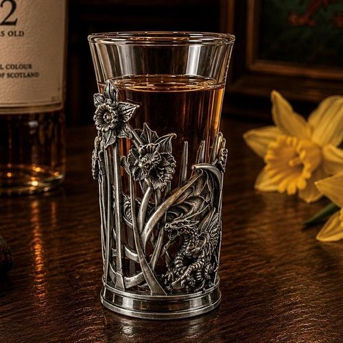A tall glass shot glass filled with amber-colored liquor, sitting on a dark wooden bar counter. The lower half of the glass is encased in a detailed pewter metal holder featuring an intricate Welsh design. The design includes textured, relief-sculpted daffodil flowers on tall stems on the left, next to a detailed mythical Welsh dragon nestled on the lower right with textured scales and wings