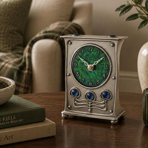 A rectangular pewter mantel clock in the style of Archibald Knox, featuring slightly flared concave sides, a stippled pewter face with rivet details, three polished blue cabochon stones set above organic sinuous metalwork, and a circular green and blue enamel dial