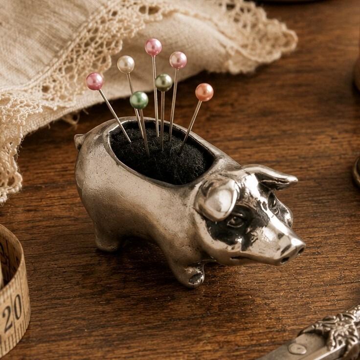 Vintage-style pewter pig pincushion figurine with a black fabric cushion on its back holding six colorful pearl-head pins