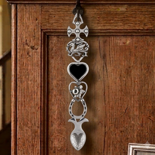 Handcrafted pewter Welsh lovespoon hanging on a wrought iron hook against a wooden panelled wall, featuring a studded Celtic cross top, heart-shaped frames enclosing a Welsh dragon, a black heart inlay, a daffodil bloom, and a lucky horseshoe above a textured spoon bowl.