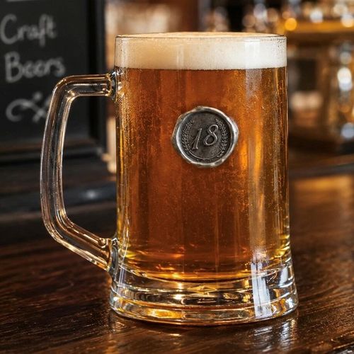 A clear glass beer tankard filled with amber ale and a thick foamy head, featuring an embossed circular silver pewter seal with the number 18 for an 18th birthday gift