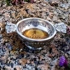 Small pewter quaich bowl filled with whiskey, engraved with the Scottish Gaelic words "Uisge Beatha" on the inner rim, displayed outdoors resting on a rugged granite rock surface