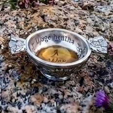 Small pewter quaich bowl filled with whiskey, engraved with the Scottish Gaelic words "Uisge Beatha" on the inner rim, displayed outdoors resting on a rugged granite rock surface