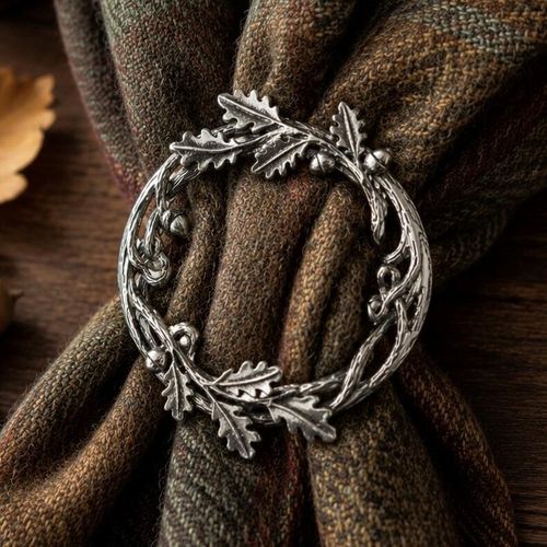 Circular pewter scarf ring styled with an intricate oak leaf and acorn wreath motif, cinching a folded green and brown tartan scarf