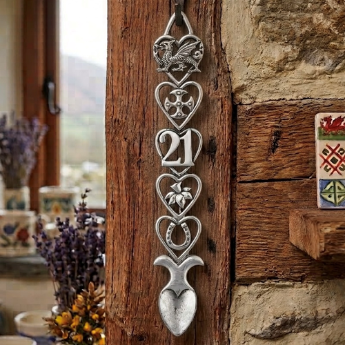 Handcrafted pewter Welsh lovespoon hanging on a rustic wooden post beside a stone wall, featuring heart-shaped frames enclosing a Welsh dragon, a Celtic cross, the number 21, a daffodil, and a lucky horseshoe above a textured spoon bowl.