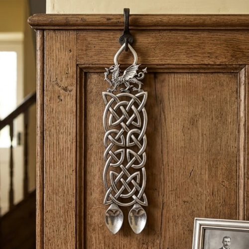 Handcrafted pewter double Welsh lovespoon hanging on a wrought iron hook against a wooden panelled wall, featuring a detailed Welsh dragon top, a broad panel of complex unending Celtic knotwork, and two side-by-side spoon bowls.