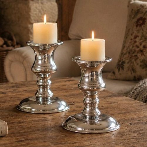 Two polished pewter candlesticks with lit ivory pillar candles displayed on a rustic wooden table in a cosy living room setting