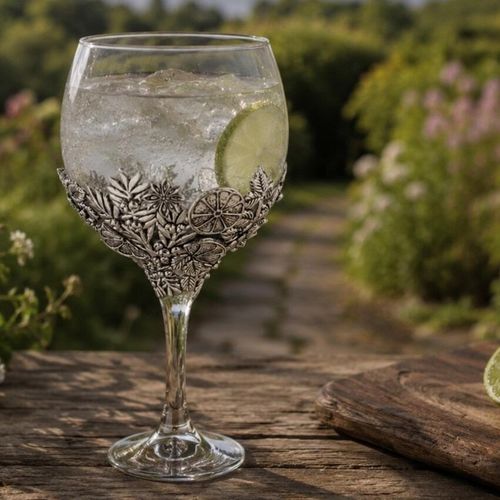 A large, balloon-shaped Copa de Balon gin glass filled with a clear, bubbling gin and tonic, ice cubes, and a fresh lime wheel. The lower portion of the glass bowl is cradled in an intricate, decorative pewter wrap detailed with sculpted botanical reliefs, including citrus slices, star anise, and leaves